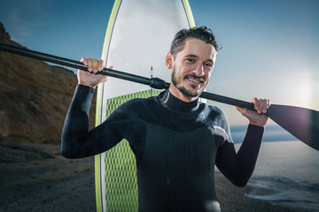 Portrait of a surfer with SUP board on the beach. Young man with a stand up paddleboard at sunset. Extreme sport concept. Male surfer lifestyle.