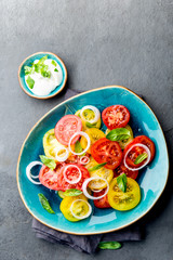 Red and yellow fresh tomato salad with onion, basil and white sauce on blue plate. Gray slate background, top view