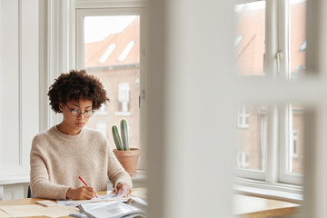 Beautiful dark skinned woman consultant rewrites information in papers, studies data, poses in spacious cabinet at home, has serious expression. Busy student prepares for examination or classes