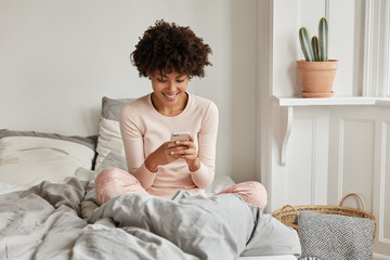 Excited delighted dark skinned housewife enjoys rest in bed, watches video via media app, has spare time, poses at comfortable bed on bedclothes, dressed in pyjamas, enjoys online marketing.