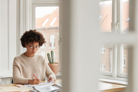 Beautiful dark skinned woman consultant rewrites information in papers, studies data, poses in spacious cabinet at home, has serious expression. Busy student prepares for examination or classes - Powered by Adobe