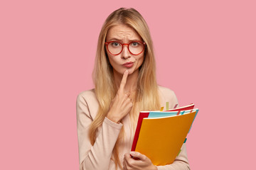 Puzzled schoolgirl has hesitant expression, purses lips, keeps index finger on chin, wears round spectacles, tries to decide what do after classes, carries books, isolated over pink background