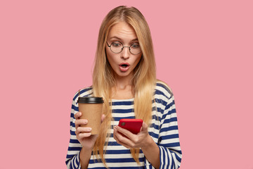 Isolated shot of surprised blonde girlfriend shocked to recieve message from lover about his attention to part, stares with terrible gaze at cell phone, carries disposable coffee, wears sailor sweater