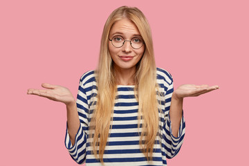 Puzzled hesitant blonde comely teenager has clueless expression, doubts about something, dressed...