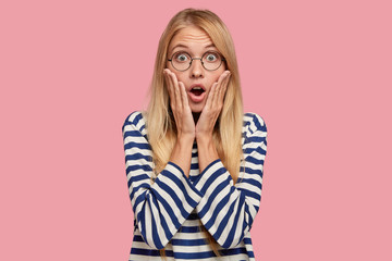 Photo of amazed blonde model keeps palms on cheeks, reacts on unexpected news from friend, stares...