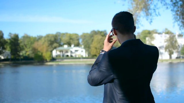 Young Businessman In A Suit And Sunglasses Is Looking At Real Estate Ofice Building By The Lake In Front Of A Beautiful Landscape