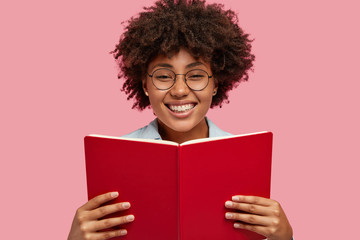Indoor shot of glad Afro American bookworm holds interesting book in front, has broad smile, happy to finish reading detective story, spends free time at home, stands over pink studio background - obrazy, fototapety, plakaty