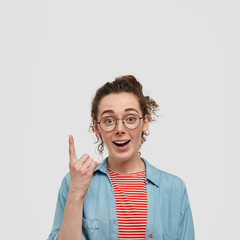 Curious surprised happy young girl has freckled skin sees something intriguing upwards, points with...