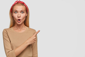 Image of surprised Caucasian woman with stupefied expression, points at upper right corner, shows blank space for your advertising content, dressed casually, stunned by something, has bated breath