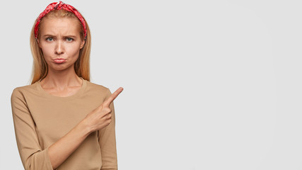 Photo of insulted attractive woman being in bad mood, points with index finger at blank copy space...