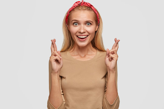 Indoor shot of attractive girl with happy expression, shining smile, light hair, crosses fingers, believes in good fortune, wears stylish red headband, beige sweater, isolated over white background