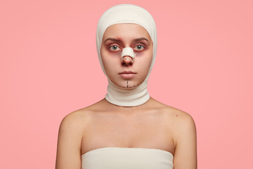 Isolated shot of girl has naked shoulders, face marked in lines, wrapped with bandage, prepared for facial treatment, going to remove wrinkles and sagging skin, isolated over pink background