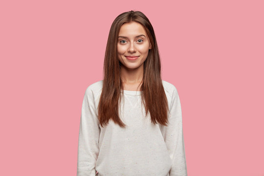 Waist up shot of attractive young girlfriend with dark straight hair soft healthy skin, dressed in casual loose sweatshirt, poses against pink background, has charming smile on face. Lifestyle concept