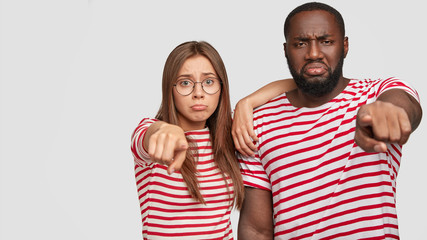 Fototapeta premium Isolated shot of dissatisfied woman and man of different nationalities, frown face in discontent, points directly at camera, dressed in casual striped t shirt, isolated over white background