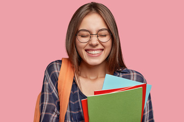 Horizontal shot of cheerful Caucasian woman with broad smile, keeps eyes closed, being in high spirit after lectures or classes, has orange rucksack, stands indoor. Positive emotions concept