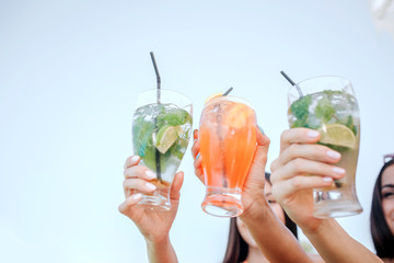 Pictire of women hold three glasses with cocktails. They show it to camera. Brunettes have rest.