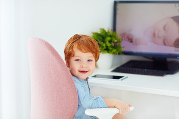 Fototapeta na wymiar happy redhead toddler baby boy is sitting in office chair at working place