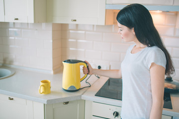 Picture of brunette stands and looks on kettle. She turns it on. Woman holds hand on the side of kettle. She is in kitchen.