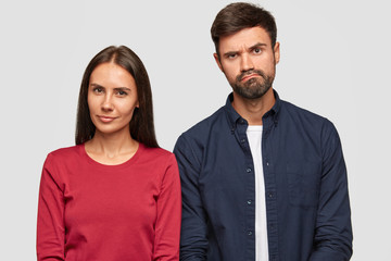 Suspicious beautiful young woman and puzzled unshaven guy stand together against white background, express different emotions, listen some information from interlocutor. Companioship concept.