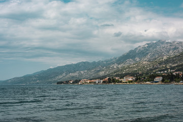 The rocky mountains in clouds from sea. Blue sea and stone coastline. The town in the distance.