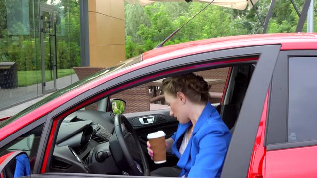 Serious business woman comes to car. She holds cup of coffee and notes. Girl openes front door and sits in car. She continues conversation on phone and make some notes. Then woman stopped talking.