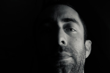 Dramatic portrait of a young man half expressionless face in deep shadow looking at camera isolated on black background