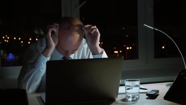 Tired businessman rubbing eyes while executive working by notebook in night office
