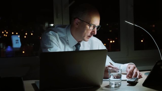 Thoughtful business man making note in notebook while last work in dark office