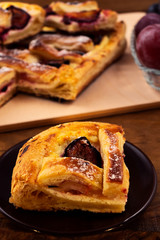 A piece of puff pastry pie with cottage cheese filling and with plums, stacked on a plate, vertical frame, wooden background.