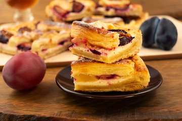 Pieces of cake with cottage cheese and plums, stacked on top of each other on a plate, next to a wooden background ripe plum purple.