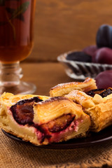 A delicious dessert is a cake of puff pastry with a cheesecake layer and baked plums, a cup of tea from the crumpled, vertical frame, wooden background.