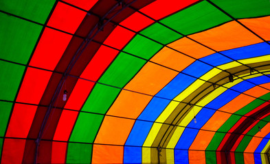 A photo taken inside a colorful tent. The tent consists of green, red, orange, blue and yellow colors and form repetitive patters of color strips. There are lamps hanging down the tent props.