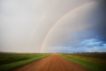 Half of a rainbow arching over a gravel road divivding green agricultural crops with a distant forest in a summer countryside landscape