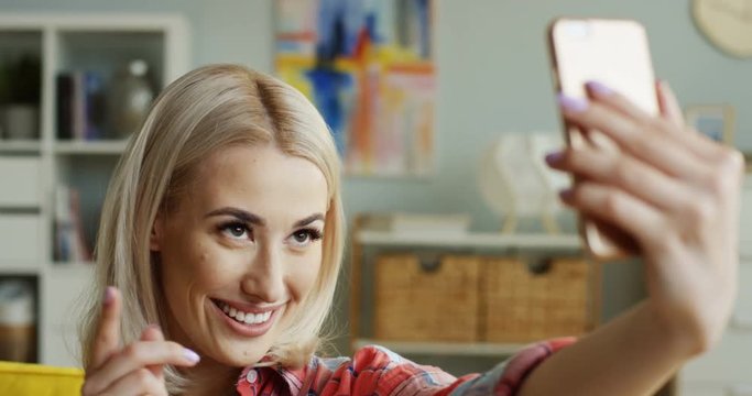 Close up of the pretty young blond woman taking selphie photos on the smartphone camera in the living room.