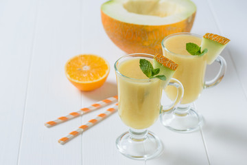 Two glass mugs with melon smoothie and fruit on a white wooden table.