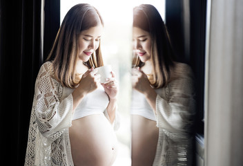 Young pregnant woman holding white cup and standing near the window