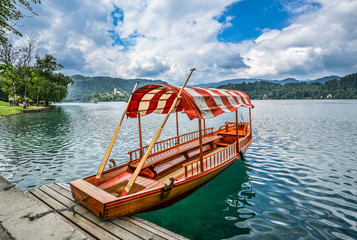 Naklejka na ściany i meble Beautiful Lake Bled in the Julian Alps and old wooden boat. Mountains, clear aquamarine water, tourist boat, lake and dramatic blue sky.