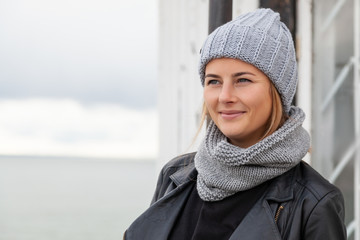 Portrait of a young woman in black coat, knitting hat, jeans walking along the pier with an old...