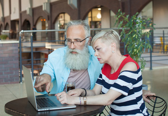 Two elderly people are sitting at cafe at the weekend, drinking coffee and watching films using the computer on the table, the senior man is pointing on the monitor