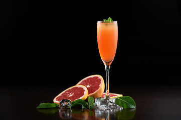 Original composition of tall cocktail glass with cold summer refreshing orange juicy drink, slices of grapefruit, fresh green mint leaves and transparent ice cubes on deep black copy space background.