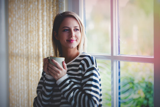 Young girl with cup of coffee stay near window at home. Autumn season