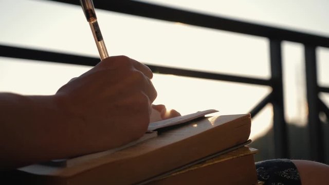 Woman's hand close-up, writing a note or letter to a notebook with a pen on a sunny day at sunset. A stack of scientific books, preparation for exams. Filling out the report