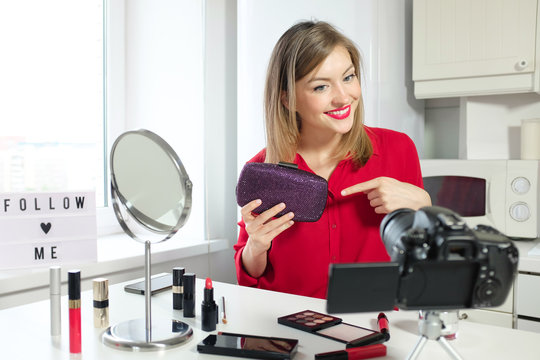 Indoor photo of young beautiful European Russian lady filming herself for beauty video blog holding purple clutch or handbag for cosmetics, showing it to camera and expressing her opinion about it