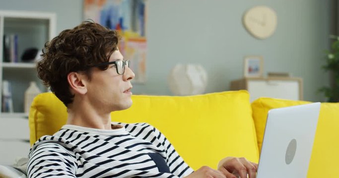 Portrait of the handsome Caucasian guy in glasses working on the laptop computer and then turning it off shutting. At home.