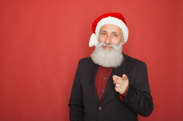 Closeup portrait of old man wearing red santa claus hat pointing with index finger in camera, isolated on red background. Positive human emotion facial expression.