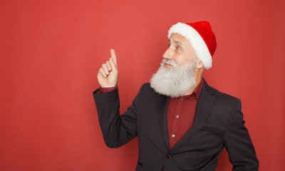 Senior man in santa hat an suit presenting something on a red wall, smiling for the camera. Xmas concept