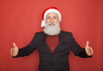 Fototapeta na wymiar Senior man in santa hat an suit presenting something on a red wall, smiling for the camera. Xmas concept