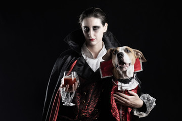 Woman and her dog in similar vampire costumes for halloween. Young female with glass of red drink and her pet puppy dressed up in same dracula costume.