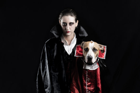 Woman And Her Dog In Similar Vampire Costumes For Halloween. Young Female And Her Pet Puppy Dressed Up In Matching Dracula Costume, Posing In Black Studio Background