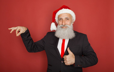 Senior man in santa hat an suit presenting something on a red wall, smiling for the camera. Xmas concept.